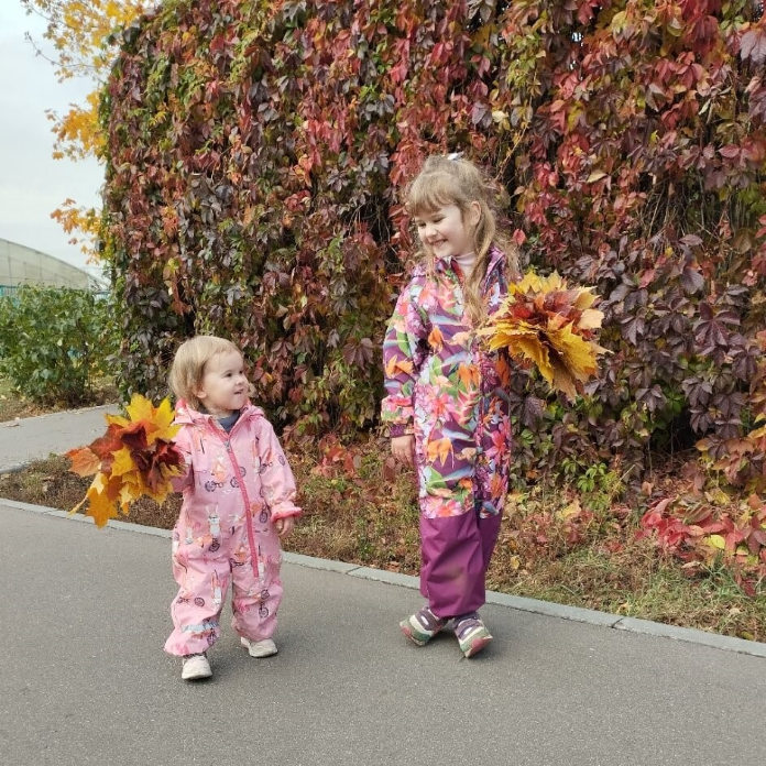 Две улыбающиеся девочки на фоне осеннего пейзажа с охапками листьев в руках.