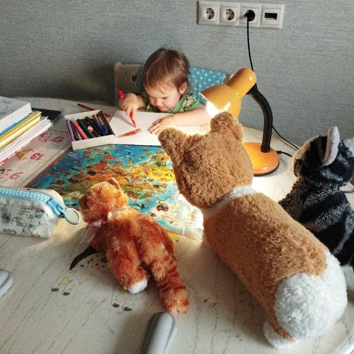 Маленькая девочка сидит на кухне за столом и старательно рисует. 
                На столе стоит включённая лампа и сидят мягкие игрушки, будто наблюдая за ней.