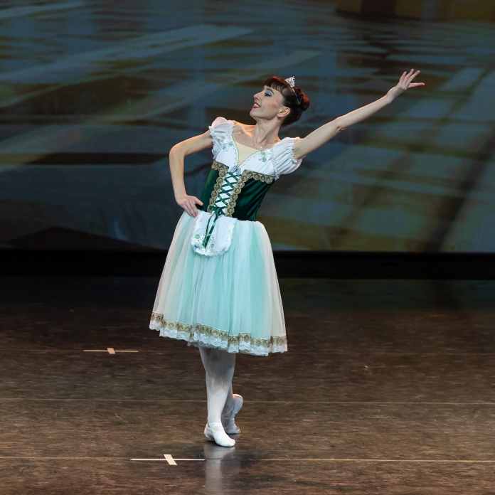 Женщина в зелено-белом балетном платье на сцене с поднятой вверх рукой. 
                Выступление - Танец невест из балета Лебединое озеро.